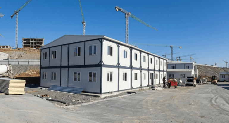 İstanbul'daki büyük ölçekli bir şantiye alanında, kule vinçler ve devam eden inşaatın önünde konumlanmış, Alyans Prefabrik üretimi iki katlı modern modüler şantiye ofis ve yatakhane binası.