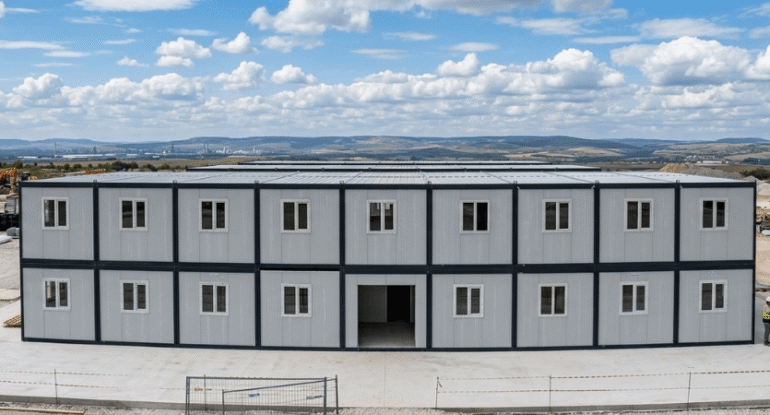 Geniş bir inşaat sahasında kurulu, gri sandviç panel duvarlı, antrasit gri karkaslı, çok pencereli ve iki katlı Alyans Prefabrik birleşimli şantiye ofis ve yatakhane binası.