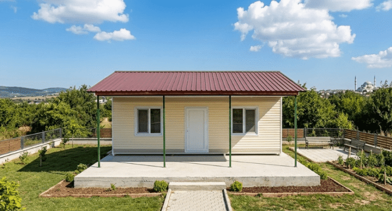 Mavi gökyüzü altında, geniş bir bahçe içerisinde konumlandırılmış; kiremit rengi çatılı, bej dış cepheli ve önünde beton verandası bulunan modern Alyans Prefabrik ev modeli. Arka planda şehir silüeti ve yeşil ağaçlar görülmektedir.